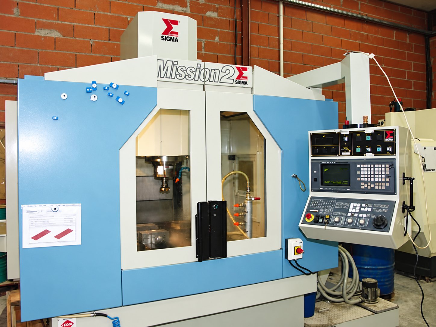Un centre d'usinage CNC bleu et blanc de marque Sigma, modèle Mission 2, installé dans un atelier aux murs de briques rouges.