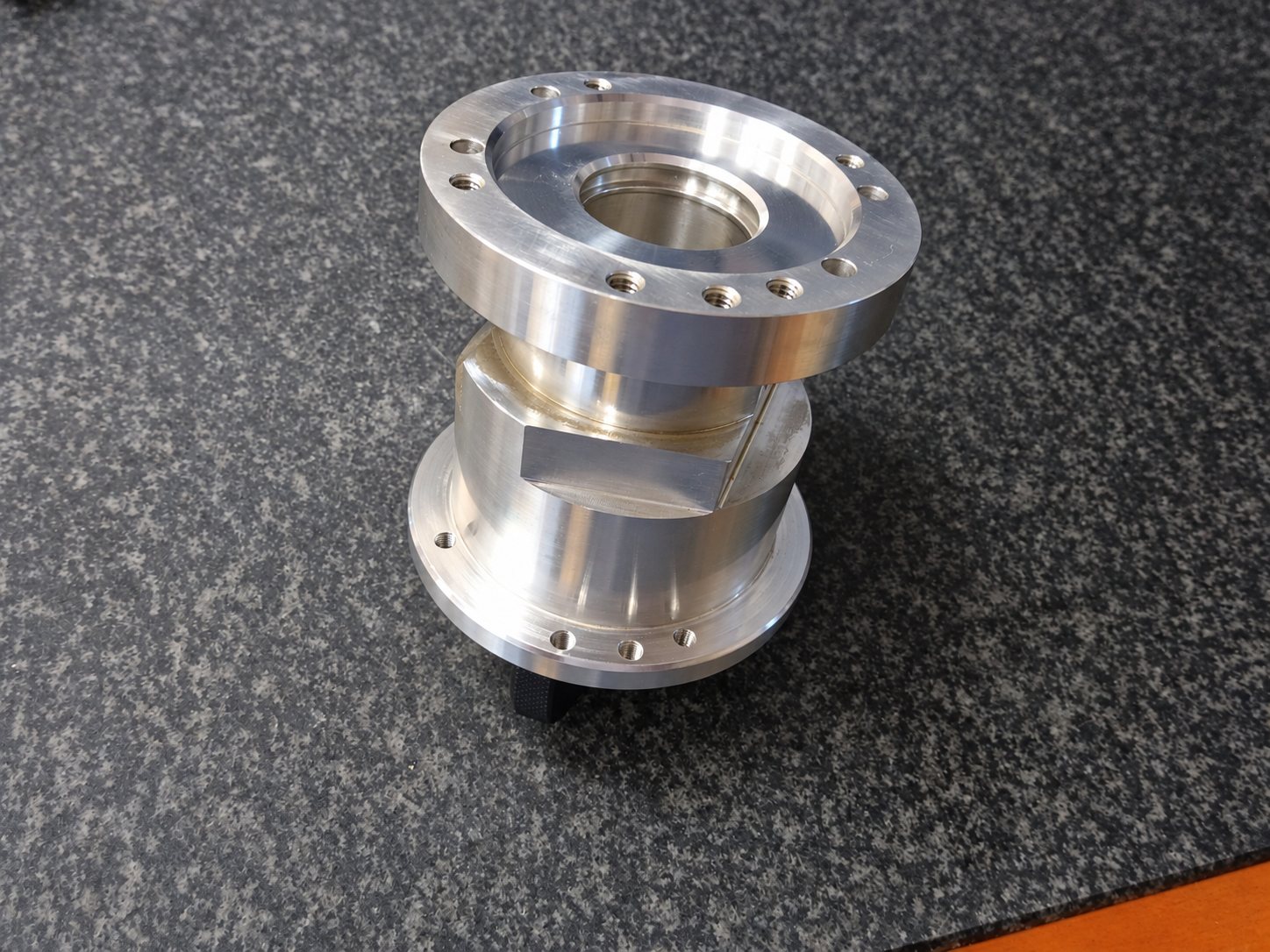Une pièce cylindrique en aluminium usiné, dotée de deux brides percées de trous, est posée sur une surface grise granitée.