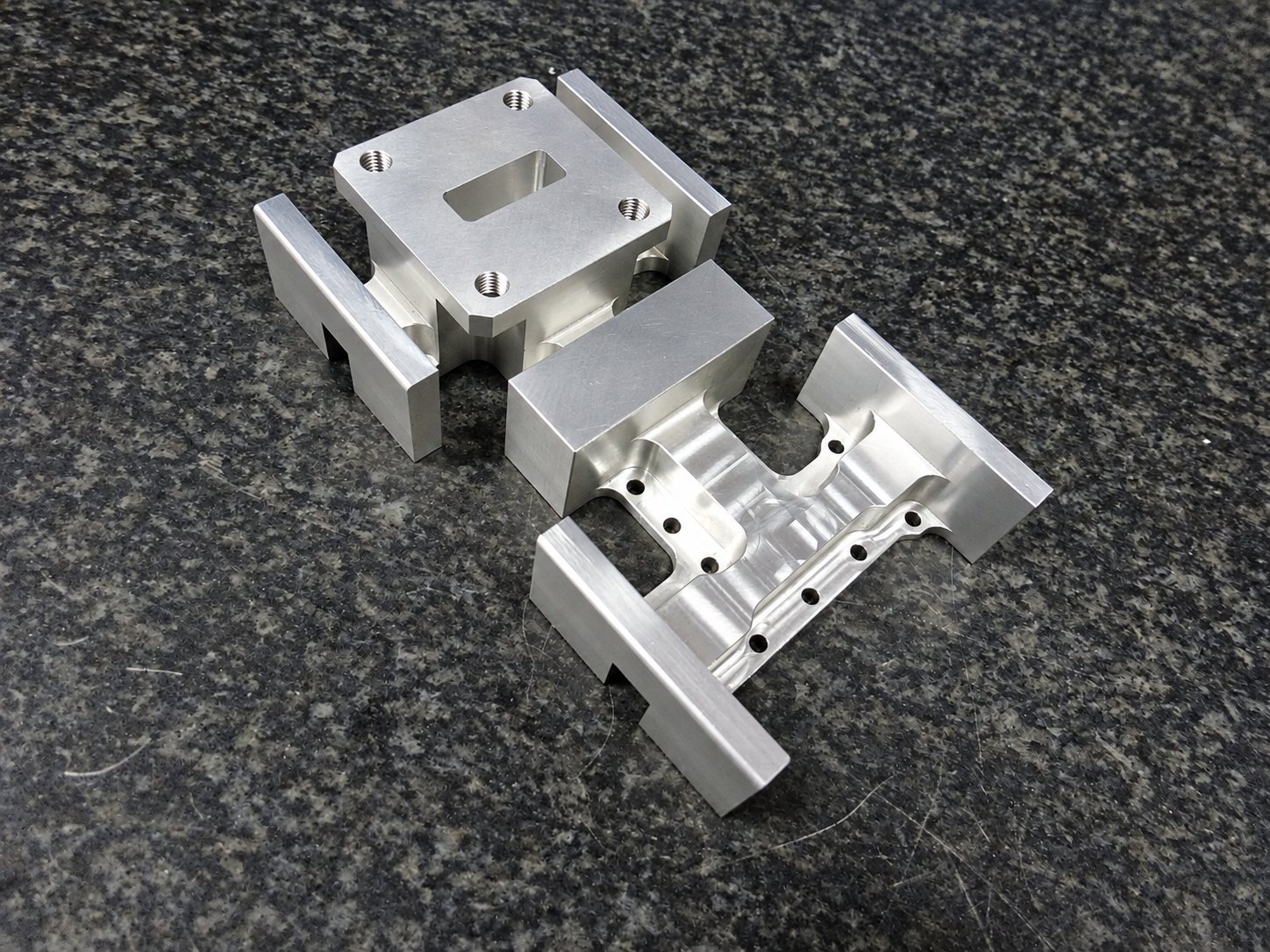 Trois pièces en aluminium usinées, percées et taraudées, posées sur une surface sombre en granit moucheté.