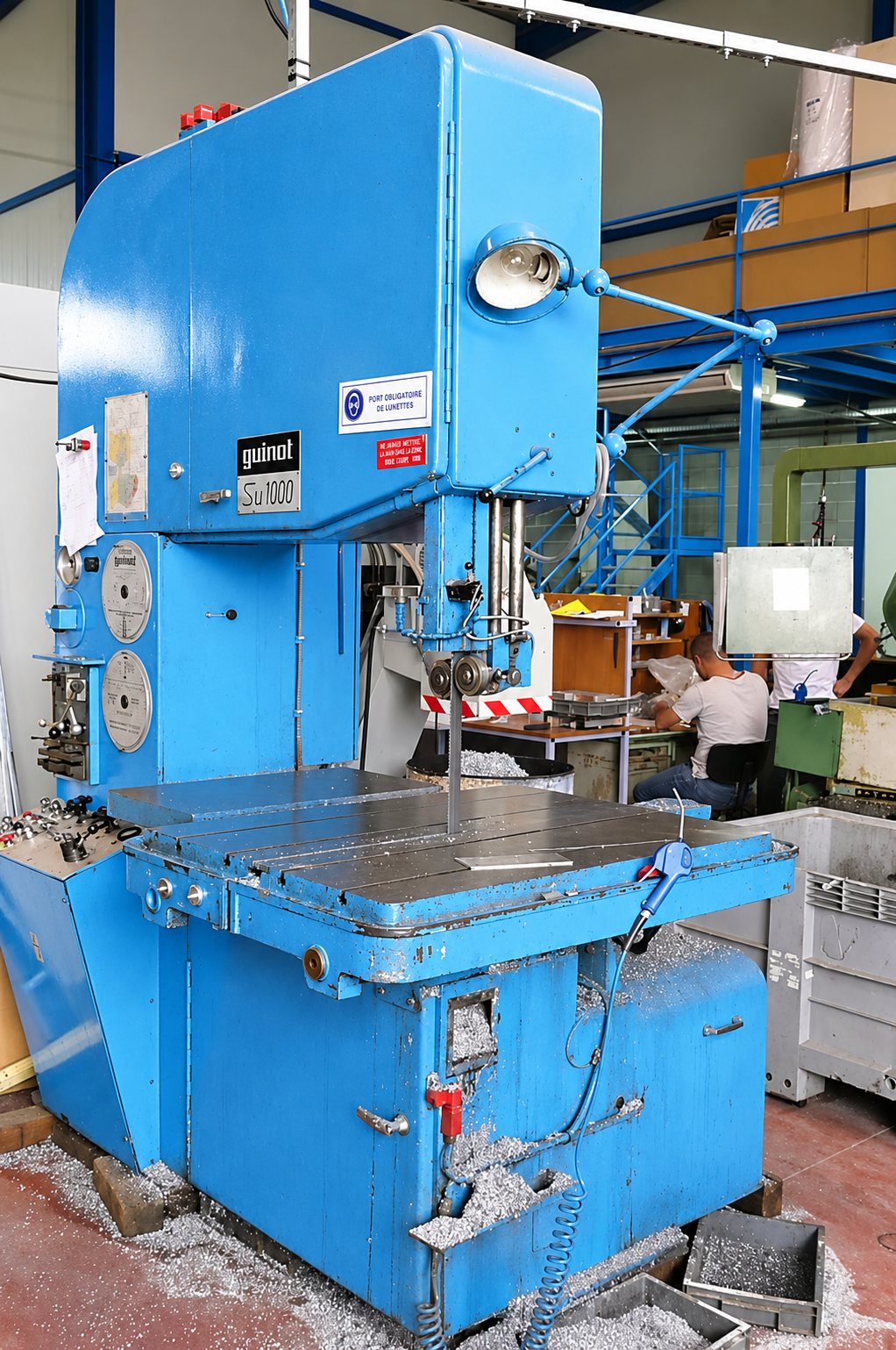 Une grande scie à ruban verticale bleue de marque Guinot, entourée de copeaux métalliques, dans un atelier où des ouvriers travaillent en arrière-plan.